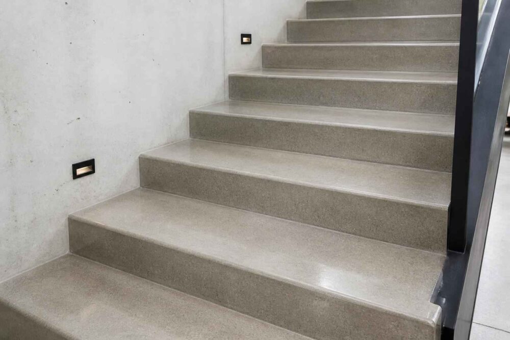 Szerokie szare schody betonowe z czarną poręczą i oświetleniem na ścianie
