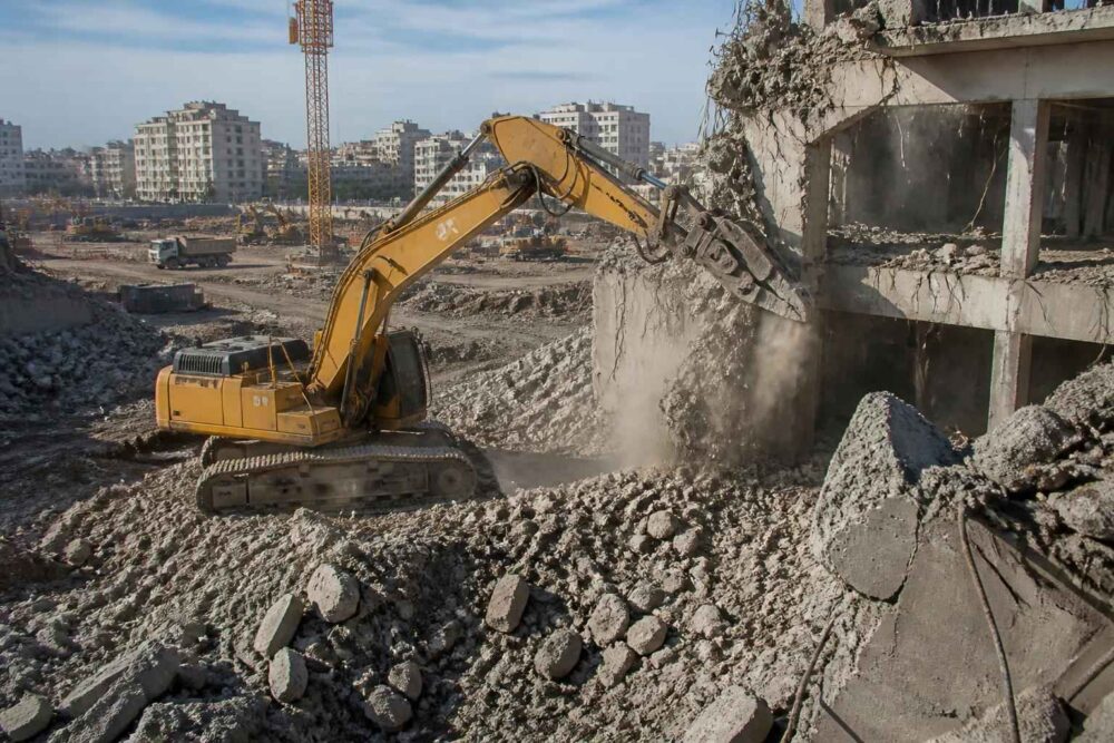 Żółta koparka kruszy betonową konstrukcję na placu rozbiórki wśród gruzów